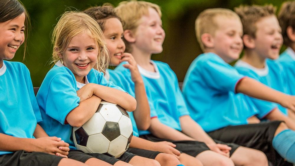  A young girl sits on a bench with a soccer ball in her hands while other children stand and sit around her.